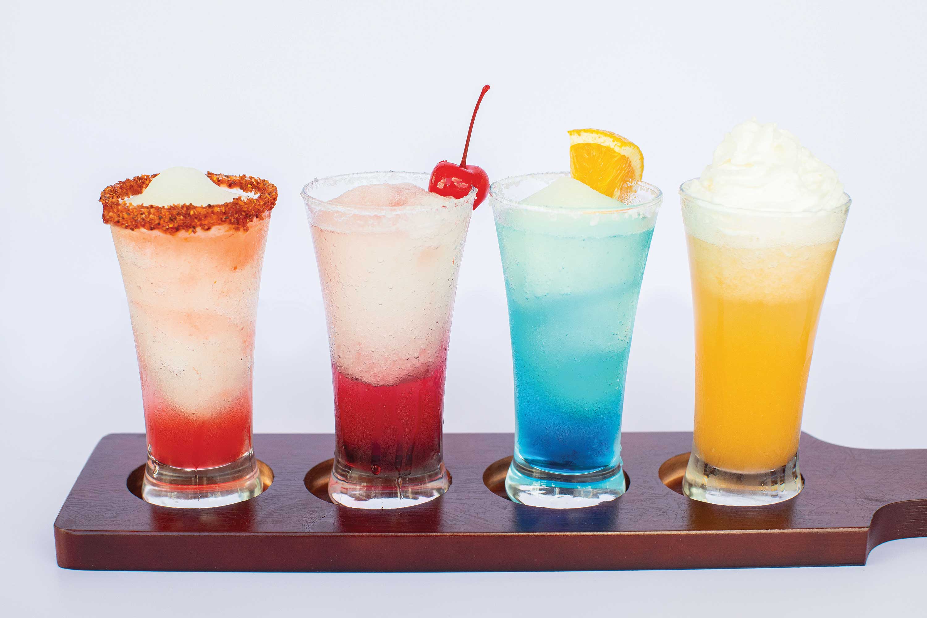 Image: Four glasses of margarita are sitting on a wooden serving platter. The first is dark pink with a salt and spice rim. The second is red with a cherry on top. The third is blue with an orange slice. The last is golden yellow with whipped cream on top.